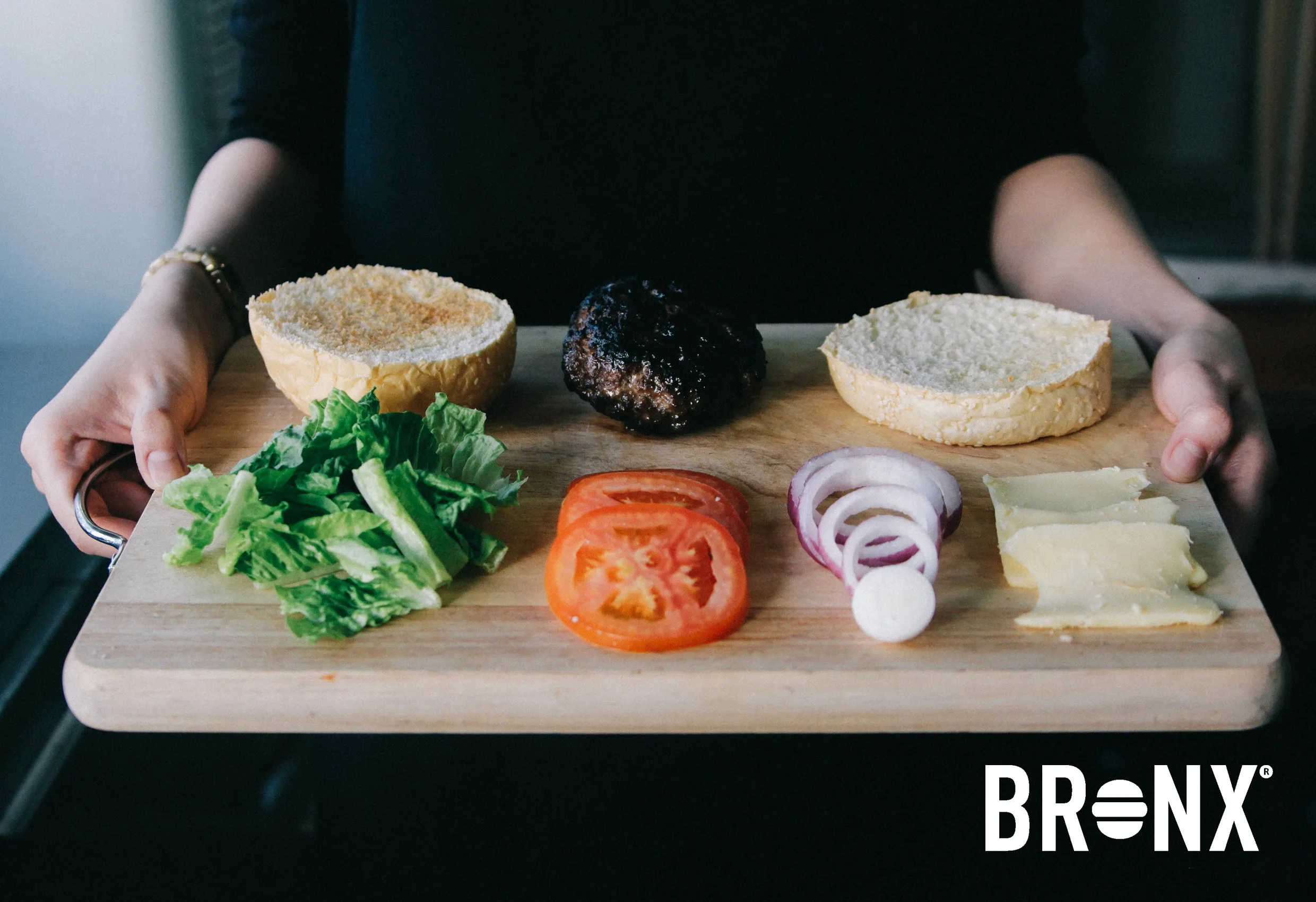 Ingredienser till en hamburgare på en skärbräda hållen av två händer, med Bronx logotyp nere till höger