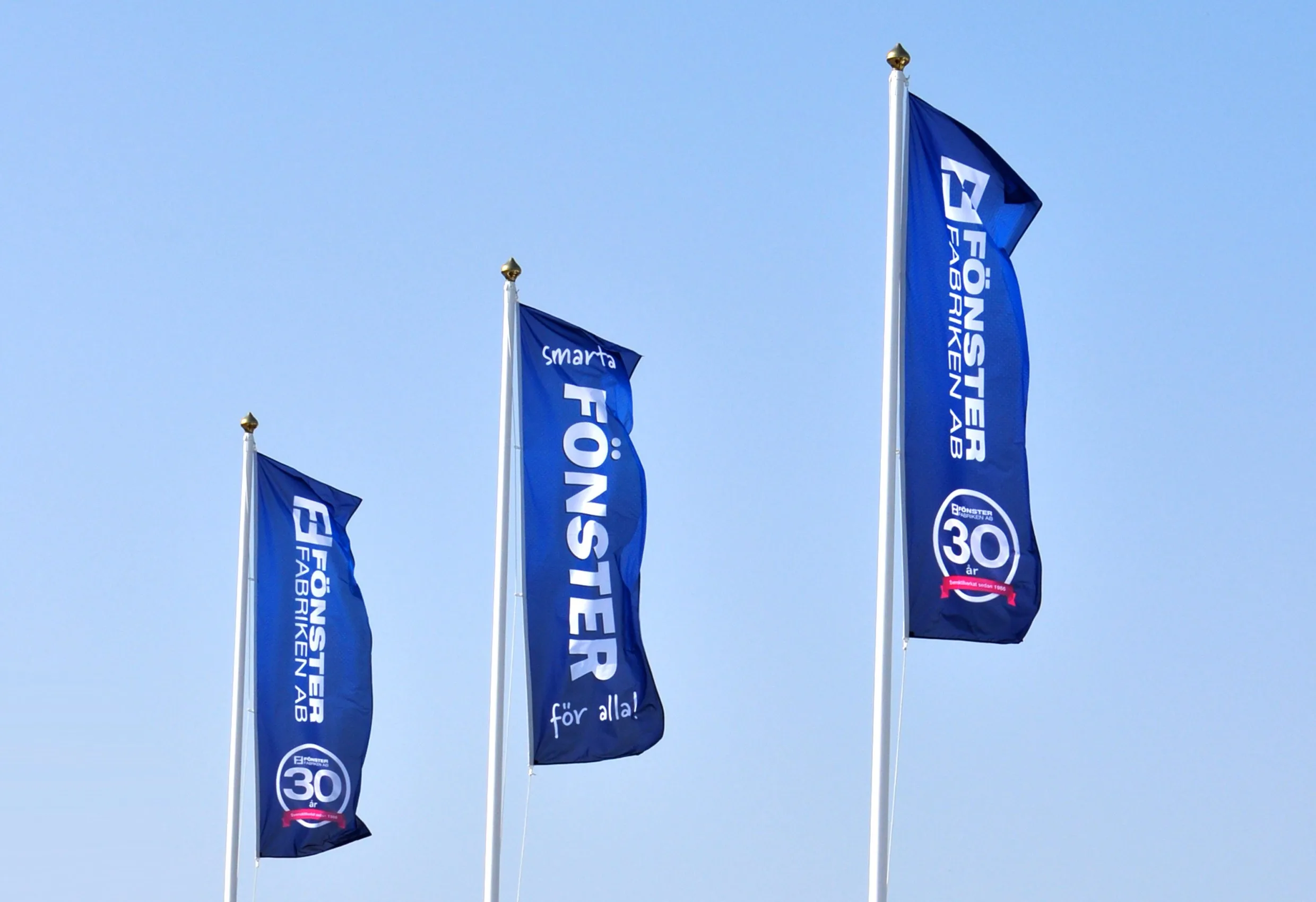 Tre blå flaggor med Fönsterfabrikens logotyp och märket ”30” mot en blå himmel