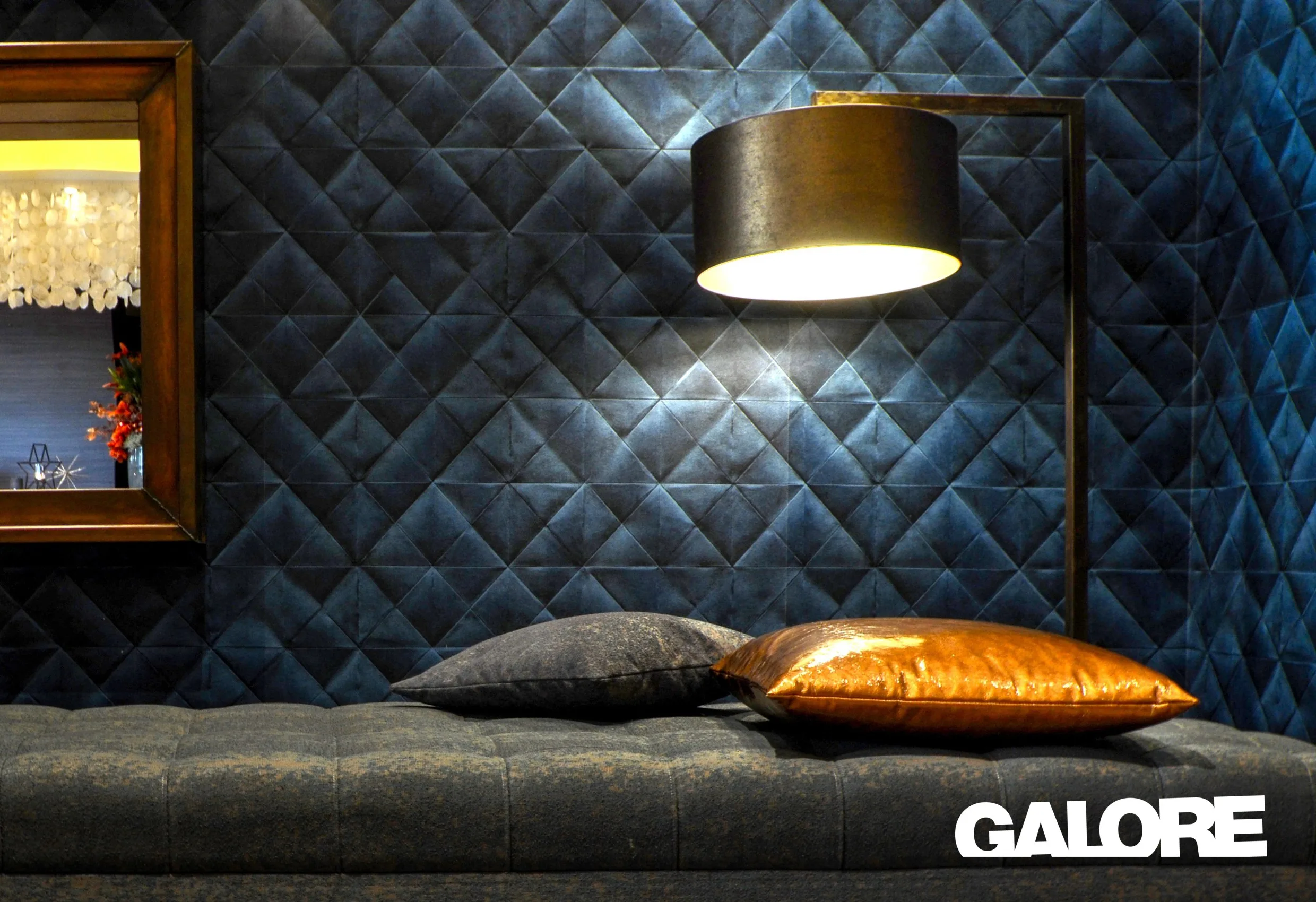 En mörkblå mönstrad vägg med en lampa och kuddar på en bänk, med Galores logotyp nere till höger