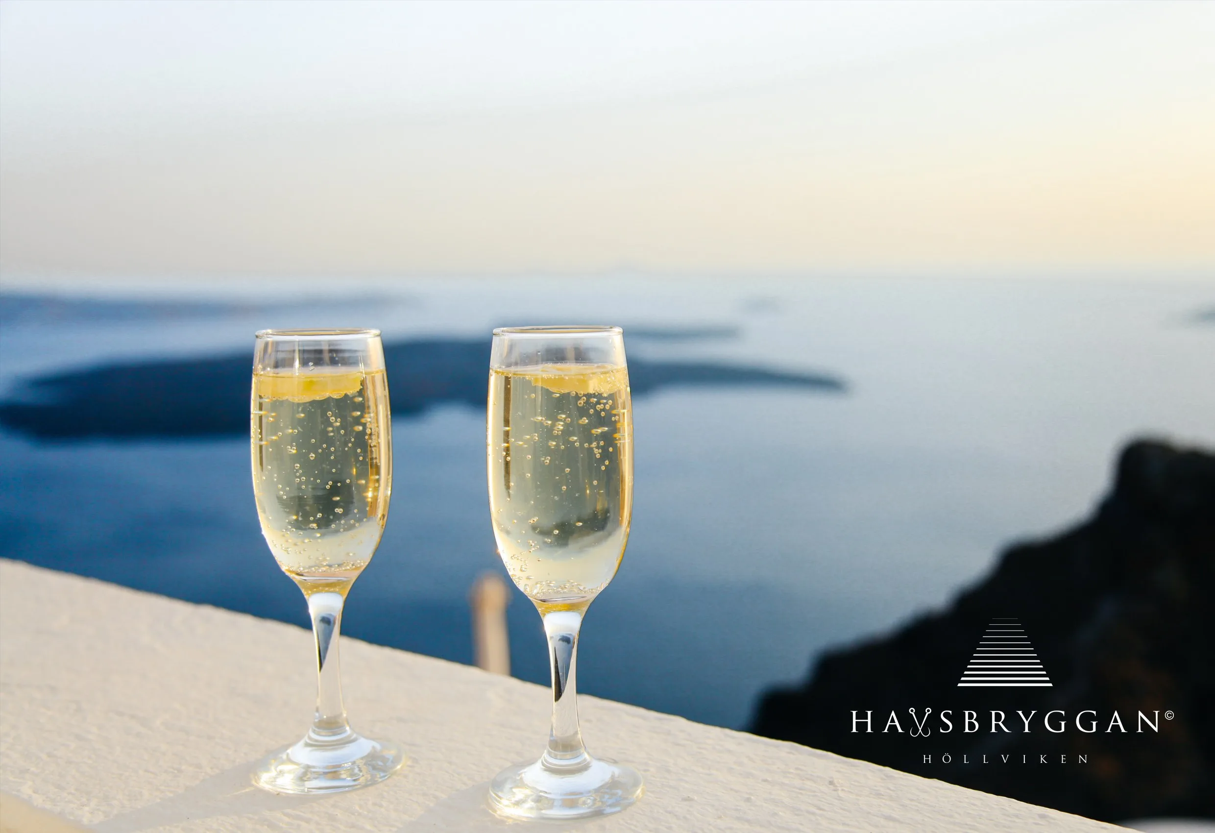 Två glas med mousserande vin vid en balustrad över havet, med Havsbryggans logotyp nere till höger