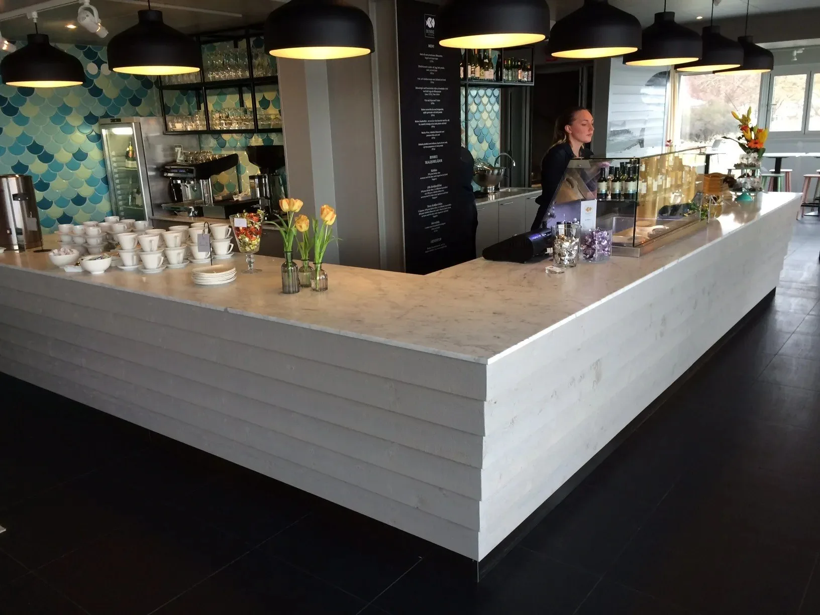 En vit bardisk i ett kafé med hängande lampor, en person bakom disken och kaffekoppar uppställda