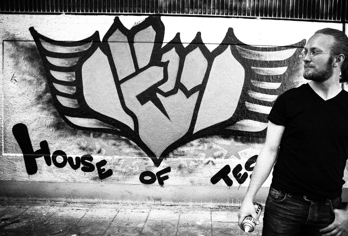 En man i svart t-shirt framför en graffitivägg med House of Tests symbol