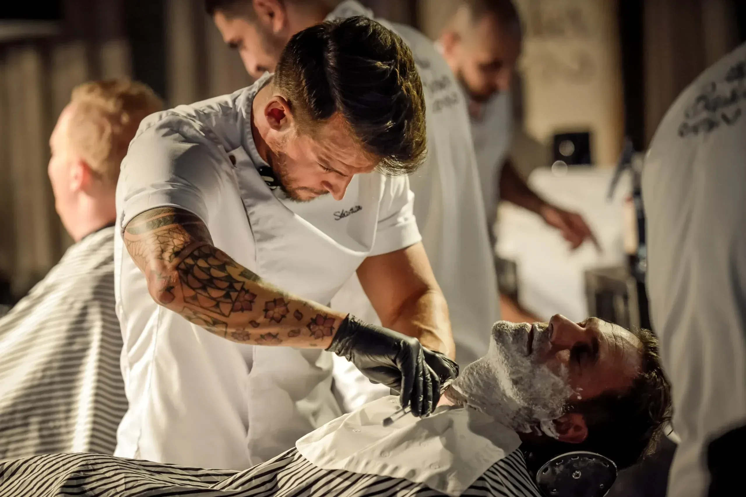 En tatuerad frisör rakar en kund på en herrfrisering, med fler personer i bakgrunden