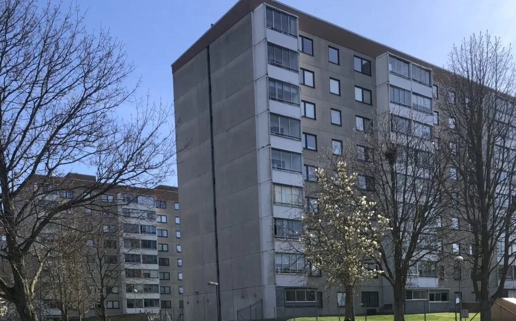 Ett högt flerbostadshus i grått med träd framför under blå himmel