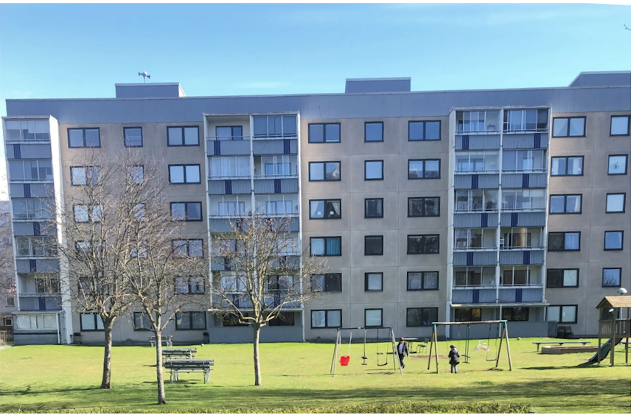 Ett långt grått flerbostadshus med balkonger och en gräsyta med lekplats framför