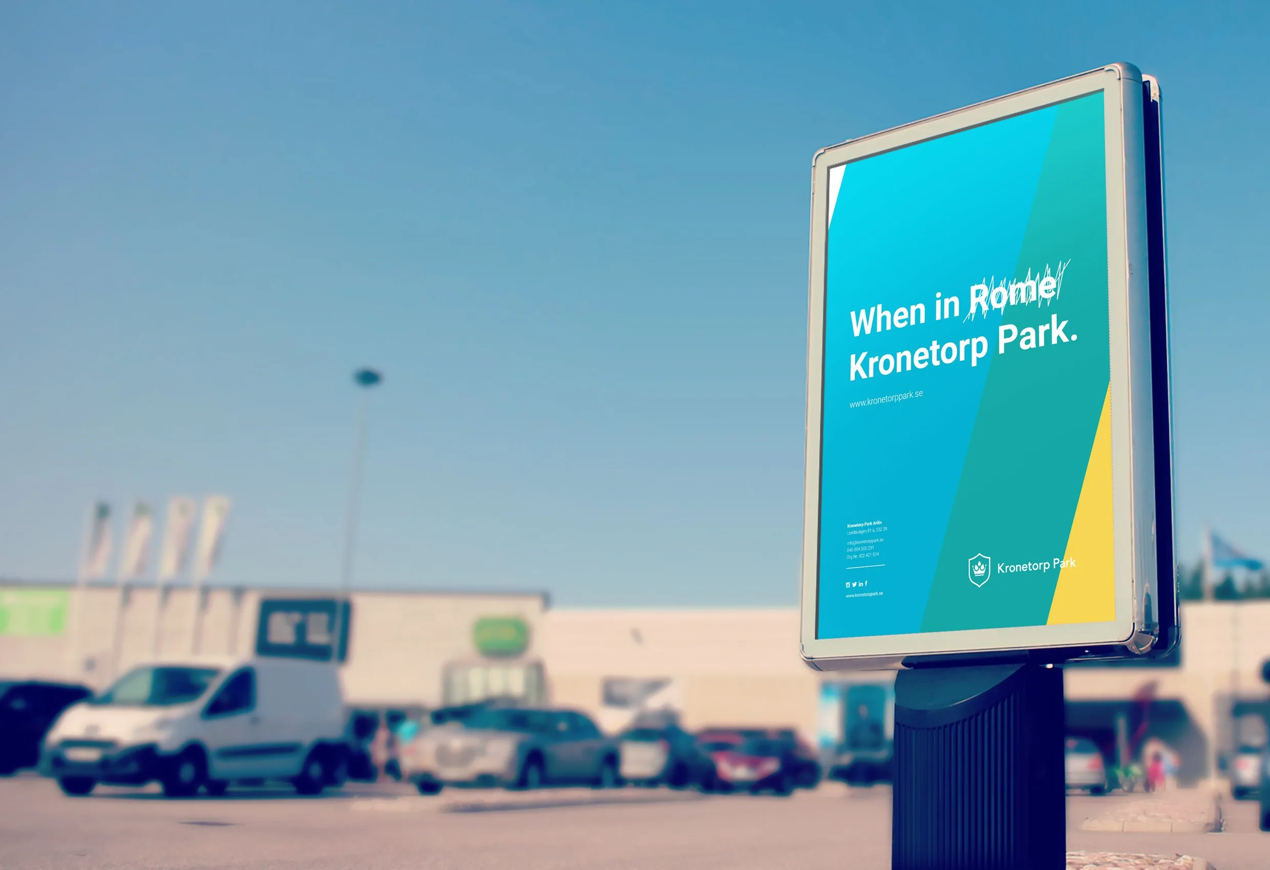 En reklampelare vid en parkering med texten ”When in Rome, Kronetorp Park.” i turkost och gult