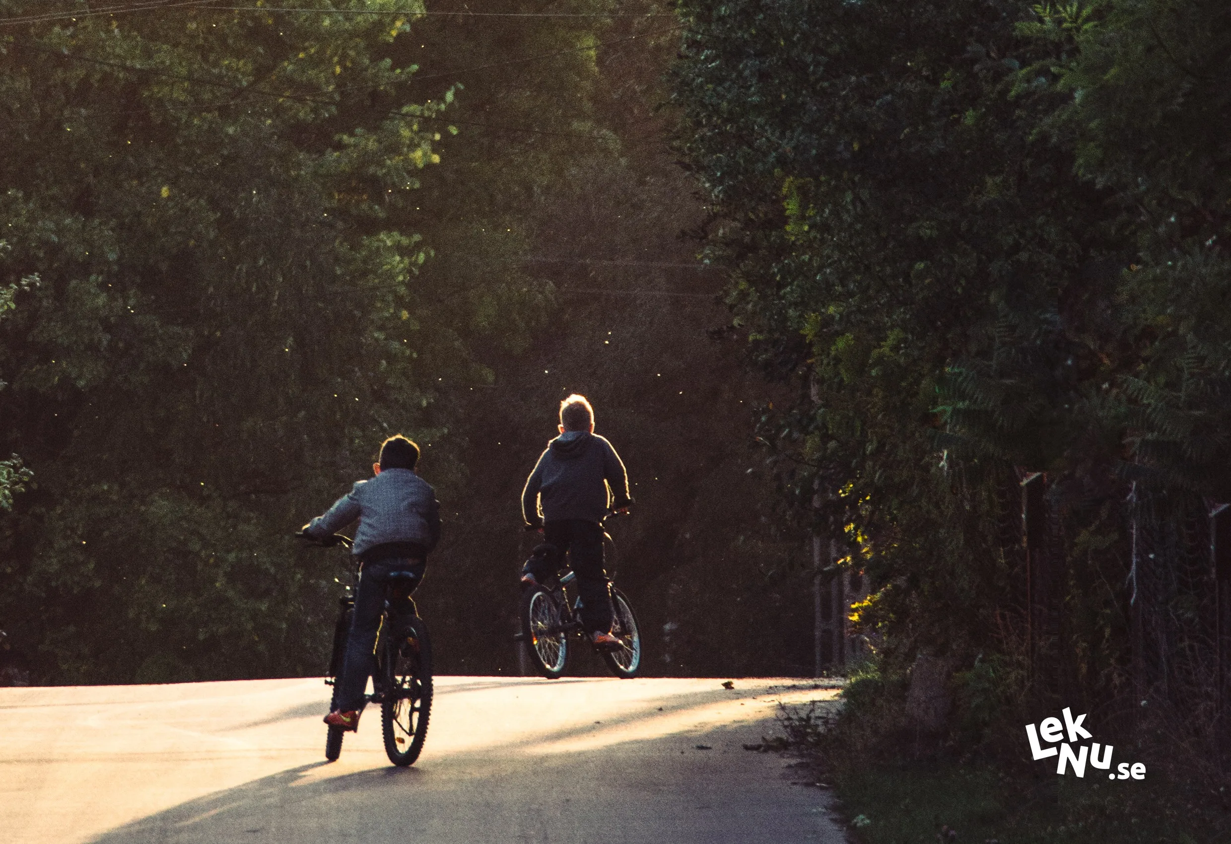 Två personer på cyklar på en skogsväg, med Lek Nus logotyp nere till höger