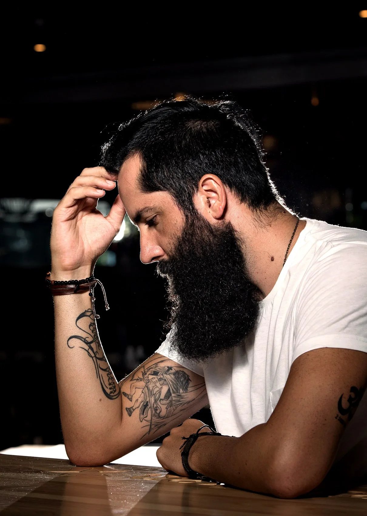 En skäggig tatuerad man i vit t-shirt som lutar huvudet i handen